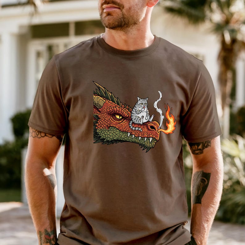 May include: Brown t-shirt featuring a cartoon dragon with red, orange, and green scales, and a gray cat sitting on its snout. The dragon is breathing fire. The shirt is a casual wear item.