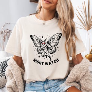 May include: Cream-colored t-shirt featuring a black and white moth-like creature with red eyes, a crescent moon, and stars. The text "NIGHT WATCH" is printed below the graphic. The shirt is a casual style.