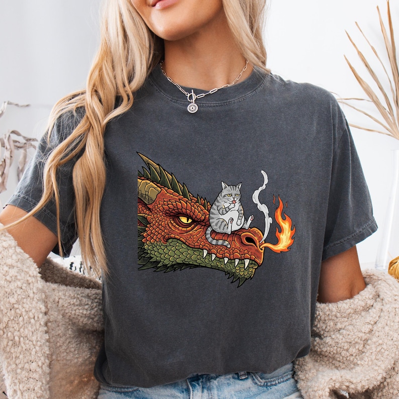 May include: Dark gray t-shirt featuring a cartoon dragon with orange and green scales, exhaling fire. A gray and white cat is perched on the dragon's snout. The shirt has a relaxed fit.