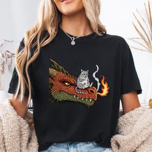 May include: Black t-shirt featuring a cartoon dragon's head with orange, red, and green scales. A gray tabby cat sits on the dragon's snout, with smoke and flames emanating from the dragon's mouth. The design is centered on the shirt.