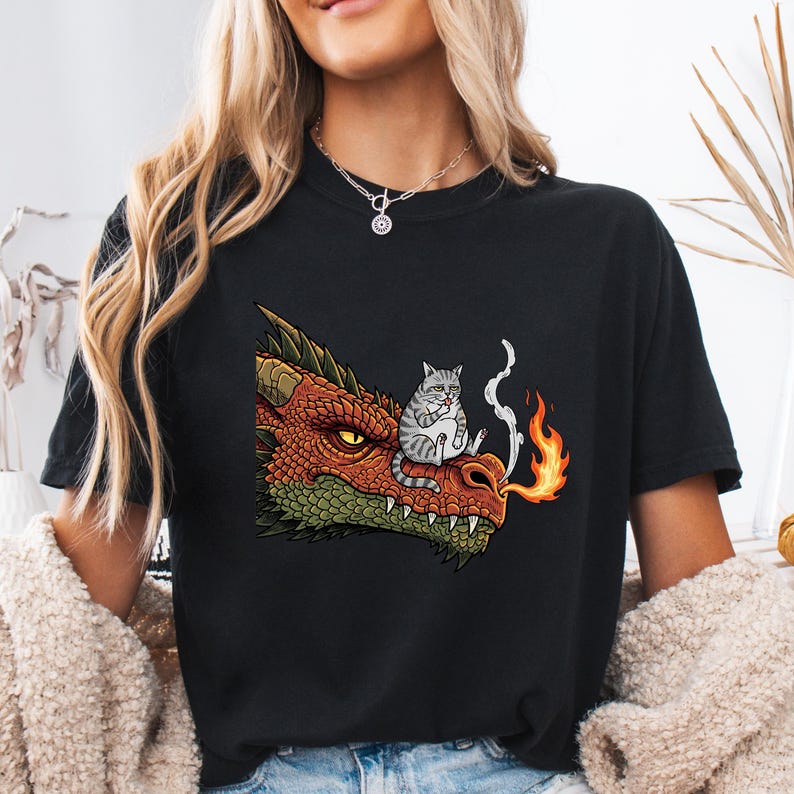 May include: Black t-shirt featuring a cartoon dragon's head with orange, red, and green scales. A gray tabby cat sits on the dragon's snout, with smoke and flames emanating from the dragon's mouth. The design is centered on the shirt.