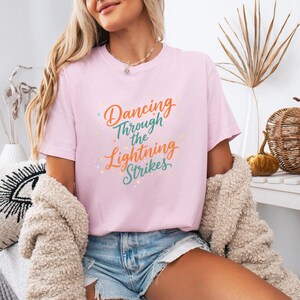 May include: A light pink t-shirt with the text "Dancing Through the Lightning Strikes" in orange, green, and yellow. The shirt is short-sleeved and appears to be made of a soft material.