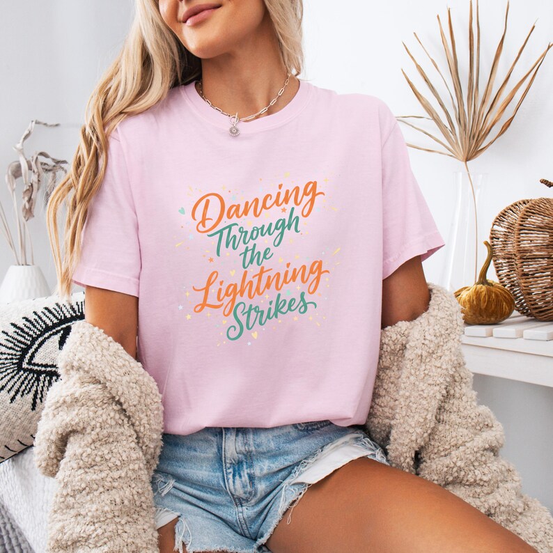 May include: A light pink t-shirt with the text "Dancing Through the Lightning Strikes" in orange, green, and yellow. The shirt is short-sleeved and appears to be made of a soft material.