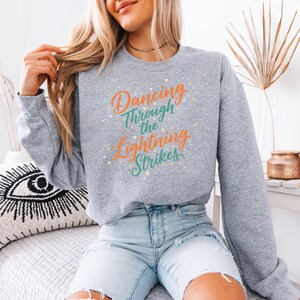 May include: A heather gray sweatshirt with the text "Dancing Through the Lightning Strikes" in orange, teal, and yellow. The sweatshirt is a crew neck style and is made of a soft material.