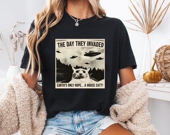 May include: Black t-shirt featuring a humorous graphic. The graphic includes the text "THE DAY THEY INVADED" above an image of UFOs and a cat, with the text "EARTH'S ONLY HOPE... A HOUSE CAT?!" below. The shirt is a casual style.