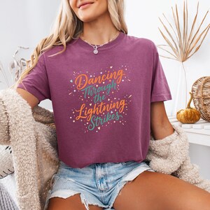 May include: A maroon t-shirt with the text "Dancing Through the Lightning Strikes" in orange, teal, and white. The shirt has a crew neck and short sleeves. The model is wearing denim shorts and a cream-colored cardigan.