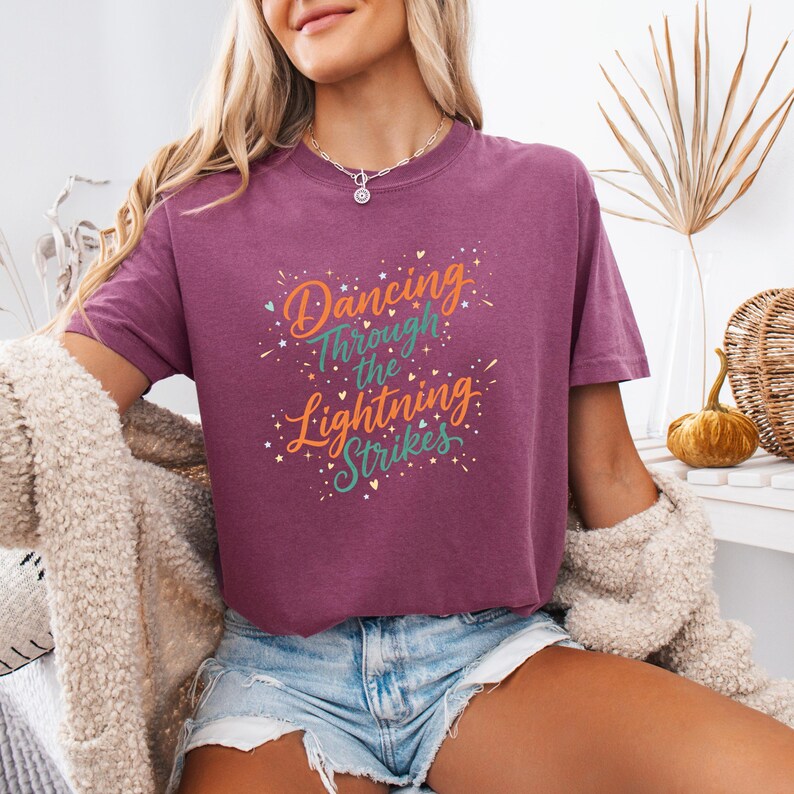 May include: A maroon t-shirt with the text "Dancing Through the Lightning Strikes" in orange, teal, and white. The shirt has a crew neck and short sleeves. The model is wearing denim shorts and a cream-colored cardigan.