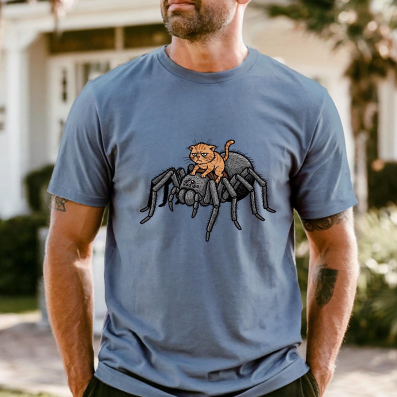 May include: A blue t-shirt featuring a cartoon illustration of an orange cat riding a large black spider. The cat has a grumpy expression. The spider has eight legs and a detailed body.