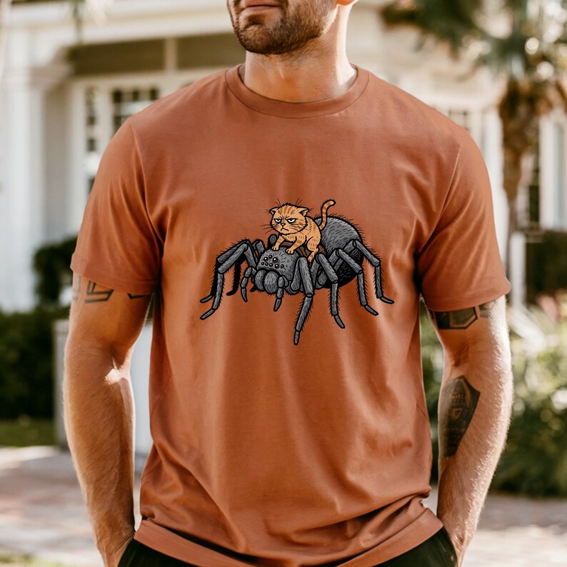 May include: Rust-colored t-shirt featuring a cartoon illustration of a gray spider with a small orange cat riding on its back. The cat has a grumpy expression. The design is centered on the front of the shirt.