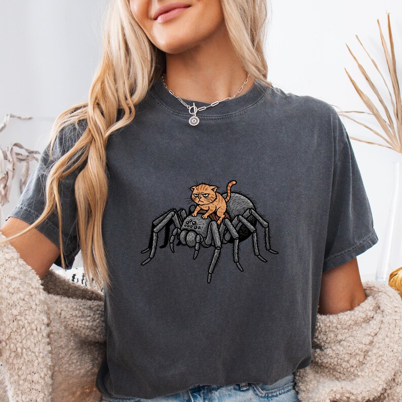 May include: Dark gray t-shirt featuring a cartoon illustration of an orange cat riding a large gray spider. The cat has an angry expression. The spider has eight legs and a detailed body. The shirt is made of a soft material.