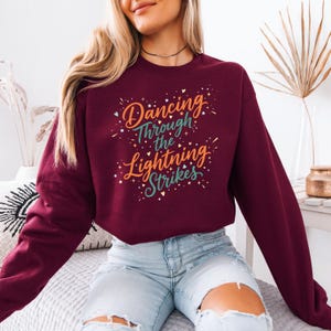 May include: A maroon sweatshirt with the text "Dancing Through the Lightning Strikes" in a colorful, decorative font. The text is surrounded by small star-like accents. The sweatshirt is a solid color and has long sleeves.