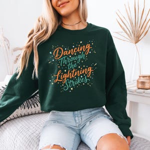 May include: A dark green sweatshirt with the text "Dancing Through the Lightning Strikes" in a colorful, whimsical font. The design includes stars and lightning bolt accents. The sweatshirt is a casual, comfortable style.