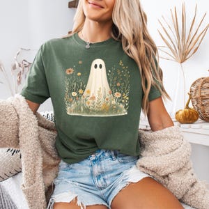 Puede incluir: Camiseta verde con un diseño de fantasma. El fantasma se muestra con una sábana blanca y ojos negros, rodeado de coloridas flores silvestres. La camiseta es de estilo casual.