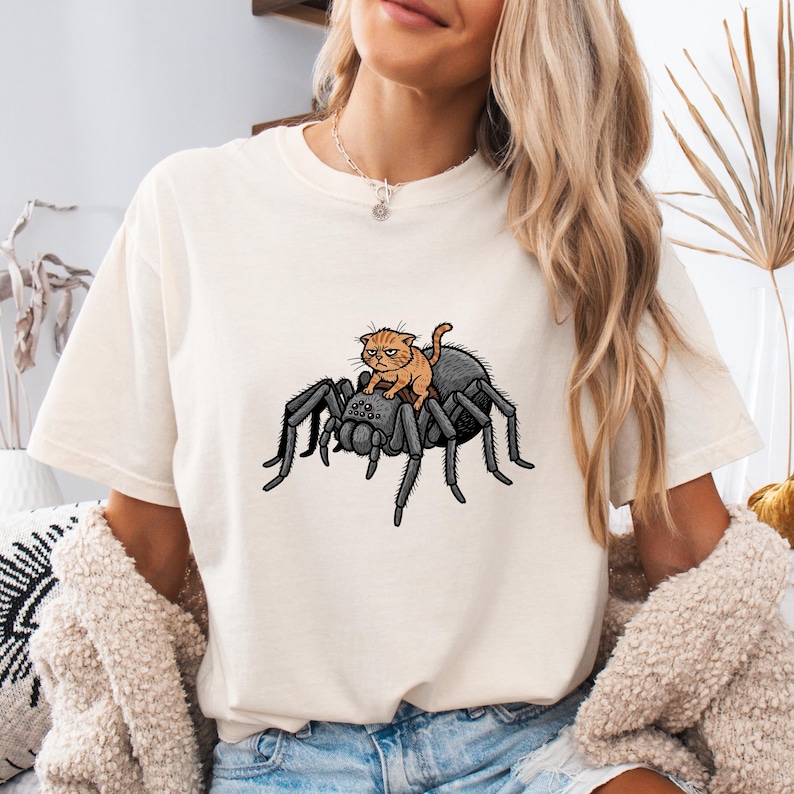 May include: Cream-colored t-shirt featuring a graphic of an orange cat riding a large black spider. The cat has a grumpy expression. The t-shirt is a casual style.