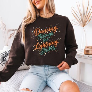 May include: A dark brown sweatshirt with the text "Dancing Through the Lightning Strikes" in orange, teal, and white. The design includes starburst accents. The sweatshirt is long-sleeved.