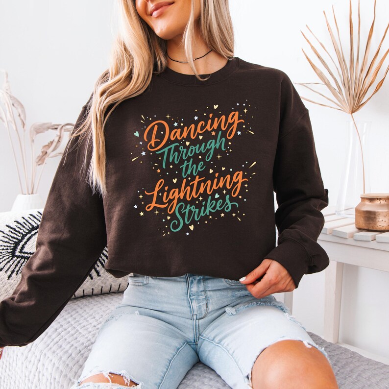 May include: A dark brown sweatshirt with the text "Dancing Through the Lightning Strikes" in orange, teal, and white. The design includes starburst accents. The sweatshirt is long-sleeved.