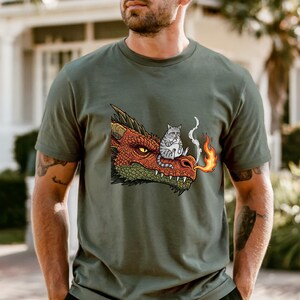 May include: Olive green t-shirt featuring a cartoon dragon's head with a cat sitting on top. The dragon is red and green, exhaling fire. The cat is gray and white. The shirt is a casual wear item.