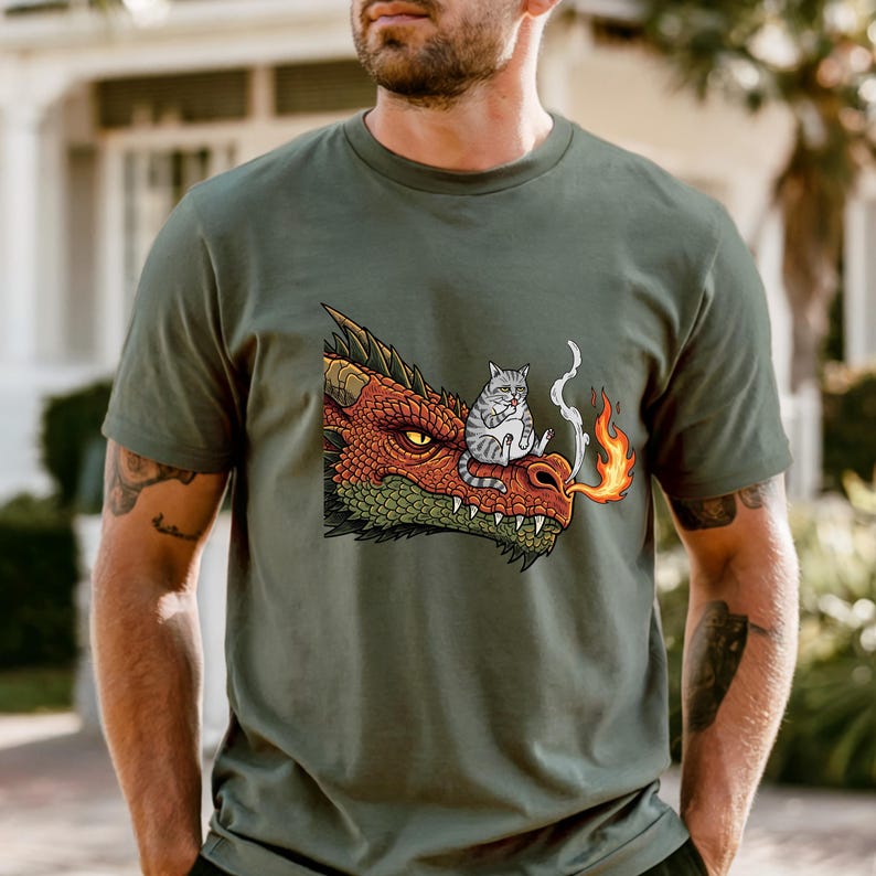 May include: Olive green t-shirt featuring a cartoon dragon's head with a cat sitting on top. The dragon is red and green, exhaling fire. The cat is gray and white. The shirt is a casual wear item.