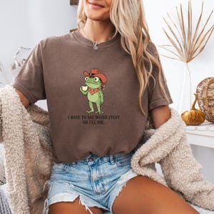 Puede incluir: Camiseta marrón con una rana de dibujos animados con sombrero de vaquero y pañuelo, sosteniendo una taza. El texto "I HAVE TO SAY WEIRD STUFF OR I'LL DIE." está impreso debajo. La camiseta la lleva una persona.