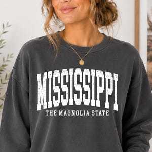 May include: A charcoal grey crewneck sweatshirt featuring the white distressed text MISSISSIPPI and THE MAGNOLIA STATE printed on the chest. The garment has long sleeves and a ribbed neckline, with the Comfort Colors logo and Pepper color label visible.