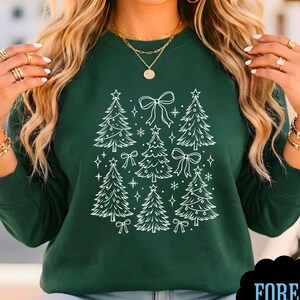 Pode incluir: Uma camisola de manga comprida em verde floresta escuro com um gráfico de linhas brancas de várias árvores de Natal, estrelas e laços dispostos num padrão central. A peça tem um decote redondo e punhos canelados nas mangas.