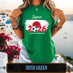 Op de afbeelding: Iers groen t-shirt met een grafisch ontwerp met het woord "Japan", een rode cirkel, een voetbal, de cijfers "2026" en de zin "GANBATTE NIPPON!". Het shirt heeft een ronde hals en korte mouwen.