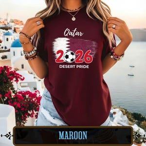Op de afbeelding: Bordeauxrood T-shirt met de tekst "Qatar 2026 Desert Pride" en een voetbalafbeelding. Het shirt heeft het ontwerp van de vlag van Qatar. Het shirt is bordeauxrood.