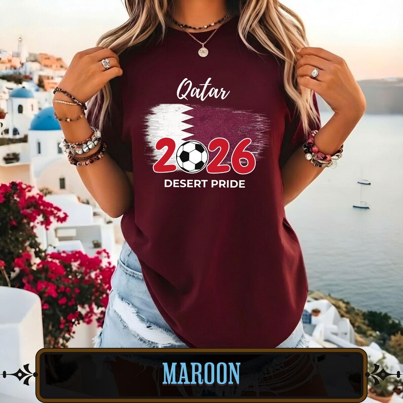 Op de afbeelding: Bordeauxrood T-shirt met de tekst "Qatar 2026 Desert Pride" en een voetbalafbeelding. Het shirt heeft het ontwerp van de vlag van Qatar. Het shirt is bordeauxrood.