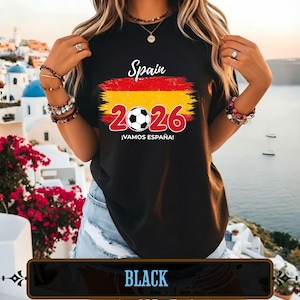 Op de afbeelding: Zwart T-shirt met een Spaans vlagontwerp en de tekst "Spain 2026 &iexcl;VAMOS ESPA&Ntilde;A!". Het ontwerp bevat een voetbal en het jaar 2026 in rood. Het woord "BLACK" staat onderaan de afbeelding.