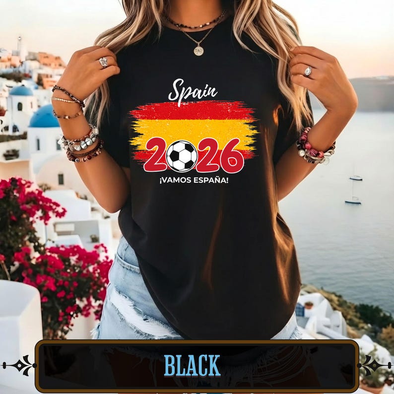 Op de afbeelding: Zwart T-shirt met een Spaans vlagontwerp en de tekst "Spain 2026 &iexcl;VAMOS ESPA&Ntilde;A!". Het ontwerp bevat een voetbal en het jaar 2026 in rood. Het woord "BLACK" staat onderaan de afbeelding.
