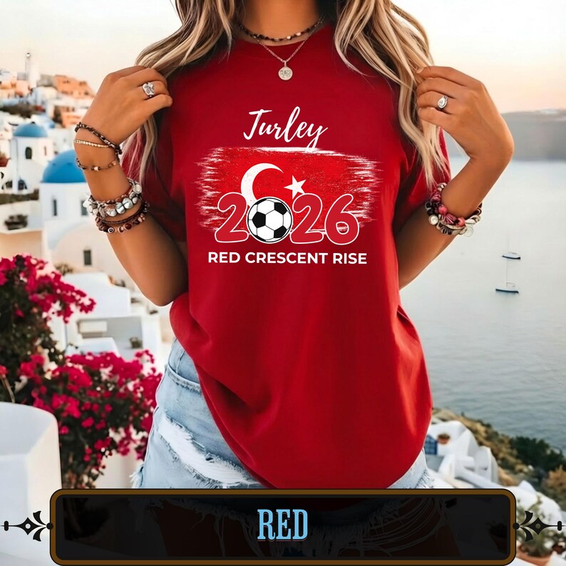 Op de afbeelding: Een rood t-shirt met het woord "Turley" in witte letters boven een afbeelding van een voetbal, een halve maan en een ster. De cijfers "2026" zijn rood en de tekst "RED CRESCENT RISE" staat eronder. Het woord "RED" staat in een zwarte doos onderaan.