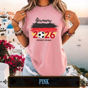 Op de afbeelding: Zachtroze T-shirt met het woord "Germany" in zwart schrift boven een afbeelding van de Duitse vlag en het nummer "2026" met een voetbal. De tekst "SCHAUT, SCHAL!" staat eronder.