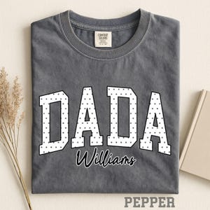 Könnte beinhalten: Ein anthrazitfarbenes T-Shirt mit Rundhalsausschnitt und dem Wort DADA in großen weißen Blockbuchstaben mit schwarzem Punktmuster. Unter dem Wort DADA ist der Name Williams in schwarzer Schreibschrift gedruckt. Das Shirt hat ein Comfort Colors Etikett.