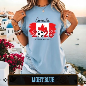 Op de afbeelding: Lichtblauw T-shirt met de tekst "Canada 2026" en een voetbalafbeelding. Het ontwerp bevat de Canadese vlag. Het shirt bevat ook de zin "We can. We will."