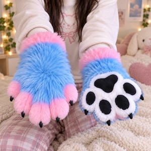 May include: A pair of plush, fuzzy paw gloves featuring bright blue fur on the back and pink fur accents. The palm side is white with black paw pads and small black claws. The wrist cuffs are trimmed with soft pink fur for a comfortable fit.