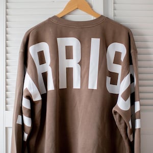 May include: A brown sweatshirt with the word "PARIS" in large white block letters across the back. The sleeves also feature white stripes. The sweatshirt is hanging on a wooden hanger against a white shuttered background.