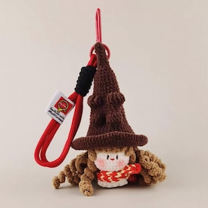 Może przedstawiać: Brelok do kluczy z szydełkową lalką czarownicy. Lalka ma brązowe włosy, brązowy kapelusz i czerwono-białą chustę. Zawieszony na czerwonej smyczy z czarnym detalem i małą etykietą.