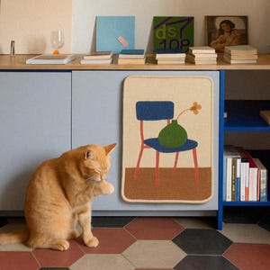 Puede incluir: Un gato atigrado naranja est&aacute; sentado cerca de un mueble con una alfombra decorativa con un dise&ntilde;o de silla, jarr&oacute;n y flor. El mueble tiene una puerta azul y una estanter&iacute;a con libros y obras de arte. El suelo tiene un patr&oacute;n geom&eacute;trico.