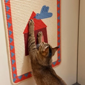 Puede incluir: Un rascador beige para gatos con un dise&ntilde;o de casa roja y nube azul. El rascador tiene un borde rojo y azul. Un gato marr&oacute;n se estira y se rasca en el rascador.
