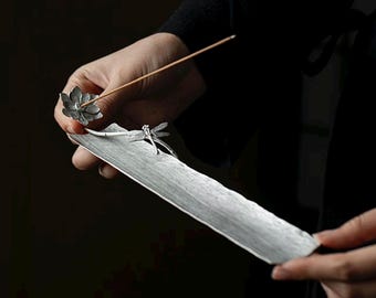 May include: A silver-colored incense holder with a lotus flower and dragonfly design. The holder is rectangular with a textured surface. An incense stick is placed in the lotus flower. The holder is held by a person.