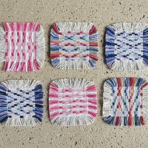 Puede incluir: Seis posavasos cuadrados tejidos con bordes con flecos. Los posavasos presentan varios patrones en tonos de rosa, azul, rojo y blanco. Los posavasos están dispuestos sobre una superficie de color neutro.