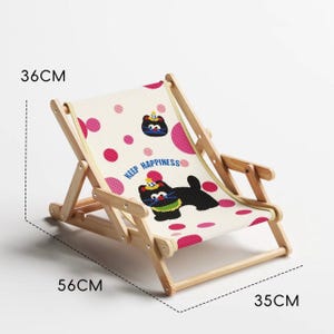 May include: A small, foldable wooden deck chair with a canvas seat. The seat is decorated with a repeating pattern of pink polka dots and cartoon dog illustrations. The chair is 14 inches high, 22 inches long, and 13.8 inches wide. The text "KEEP HAPPINESS" is printed on the seat.