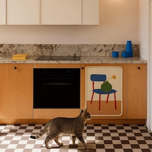 Puede incluir: Una cocina moderna con gabinetes de madera clara, un horno negro y un suelo a cuadros. Un tapiz decorativo muestra una silla y un jarr&oacute;n azules. Un gato atigrado gris camina por el suelo.