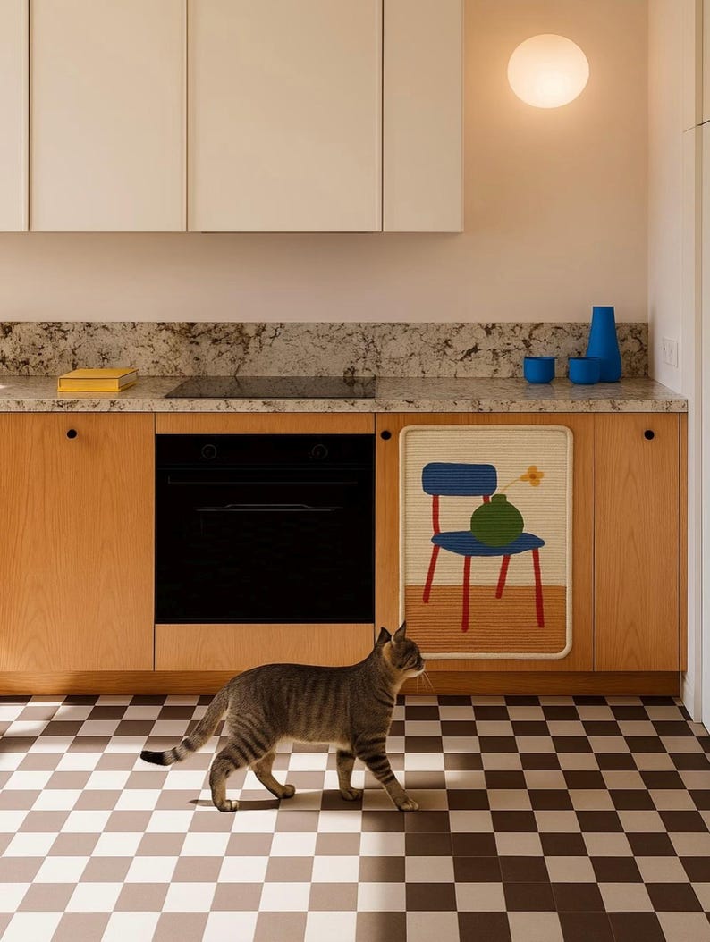 Puede incluir: Una cocina moderna con gabinetes de madera clara, un horno negro y un suelo a cuadros. Un tapiz decorativo muestra una silla y un jarr&oacute;n azules. Un gato atigrado gris camina por el suelo.
