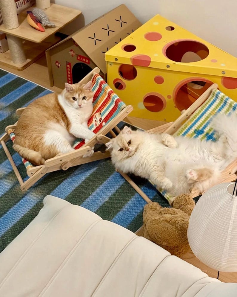 May include: Two cats lounging on miniature wooden beach chairs with colorful striped fabric. A yellow, cheese-shaped cat house and a small cardboard house are in the background. The cats are light orange and white, and cream-colored.