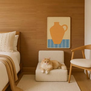 Puede incluir: Un gato de color naranja claro descansa sobre una peque&ntilde;a silla tapizada de color crema. Una obra de arte enmarcada con un dise&ntilde;o de jarr&oacute;n naranja cuelga de una pared con paneles de madera. Se ve una cama con una manta y una almohada de color crema.