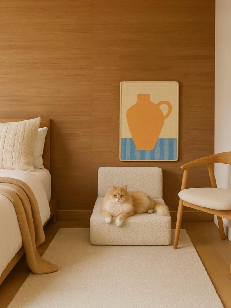 Puede incluir: Un gato de color naranja claro descansa sobre una peque&ntilde;a silla tapizada de color crema. Una obra de arte enmarcada con un dise&ntilde;o de jarr&oacute;n naranja cuelga de una pared con paneles de madera. Se ve una cama con una manta y una almohada de color crema.