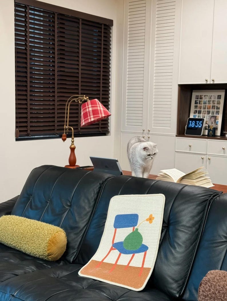Puede incluir: Un sof&aacute; de cuero negro con un coj&iacute;n decorativo y una alfombra estampada con un dise&ntilde;o de silla y jarr&oacute;n. Una l&aacute;mpara dorada y a cuadros rojos se encuentra sobre una mesa de madera. Un gato gris est&aacute; sobre la mesa.
