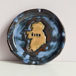 May include: A round ceramic tray featuring a glossy glaze with swirling shades of dark blue, black, and brown. In the centre, a raised, tan-coloured relief map of Ireland is displayed with the word ÉIRE embossed in black lettering on its surface.