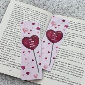 May include: Two bookmarks with a pink background, featuring heart-shaped lollipops and small hearts. The lollipops have the text "I'M A SUCKER FOR BOOKS" in a dark red colour. The bookmarks are next to an open book.